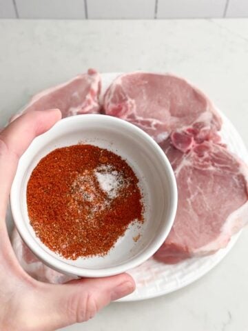 hand holding spice mix in front of plate of raw pork chops.
