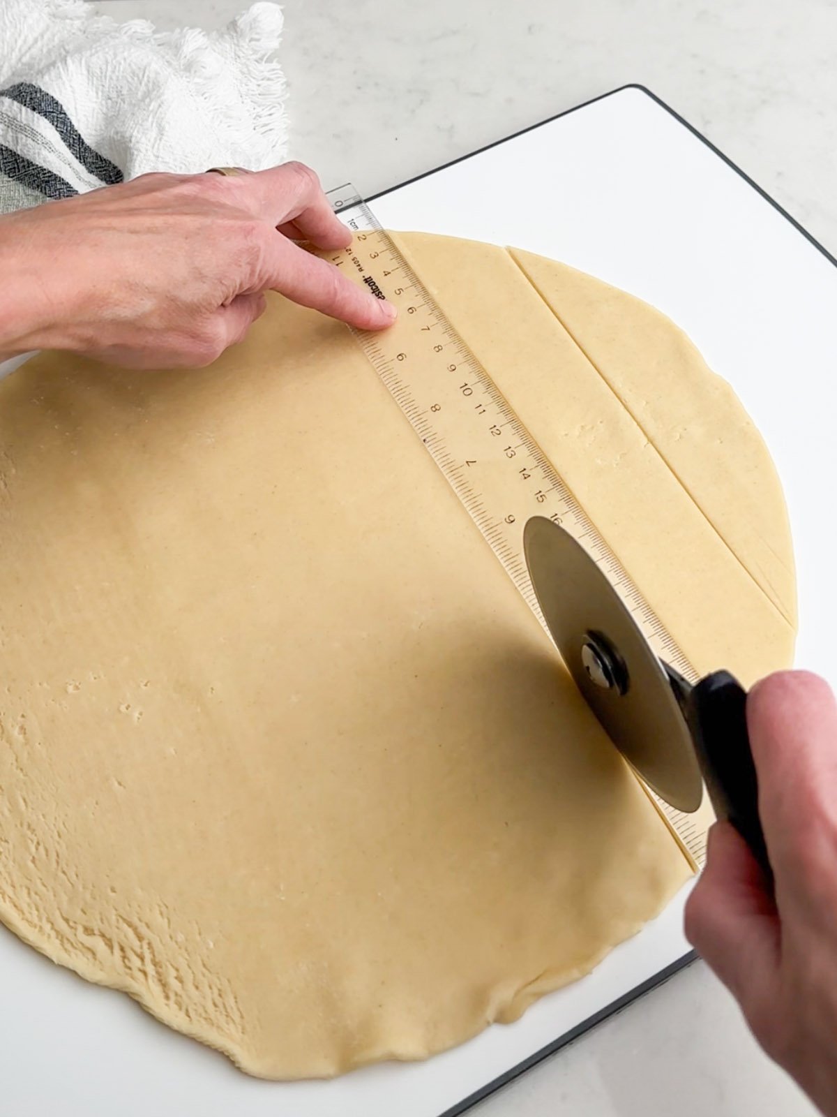 Hands holding a pizza cutter and ruler cutting pie dough into strips.