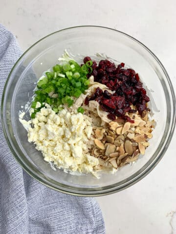 Chicken, cranberries, green onions, feta, and almonds in a clear bowl. 