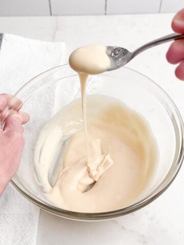 hand holding a spoon with icing drizzling down into a clear bowl of icing.