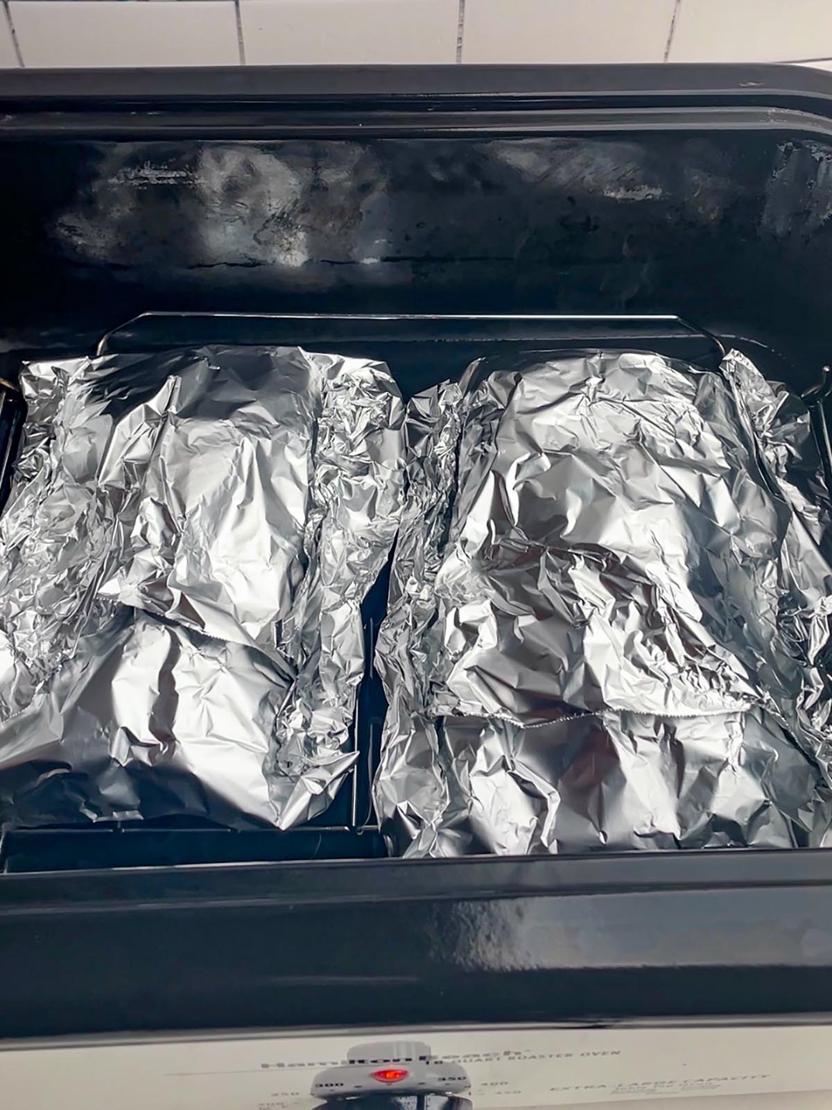 two foil packets of ribs in a black roaster oven pan