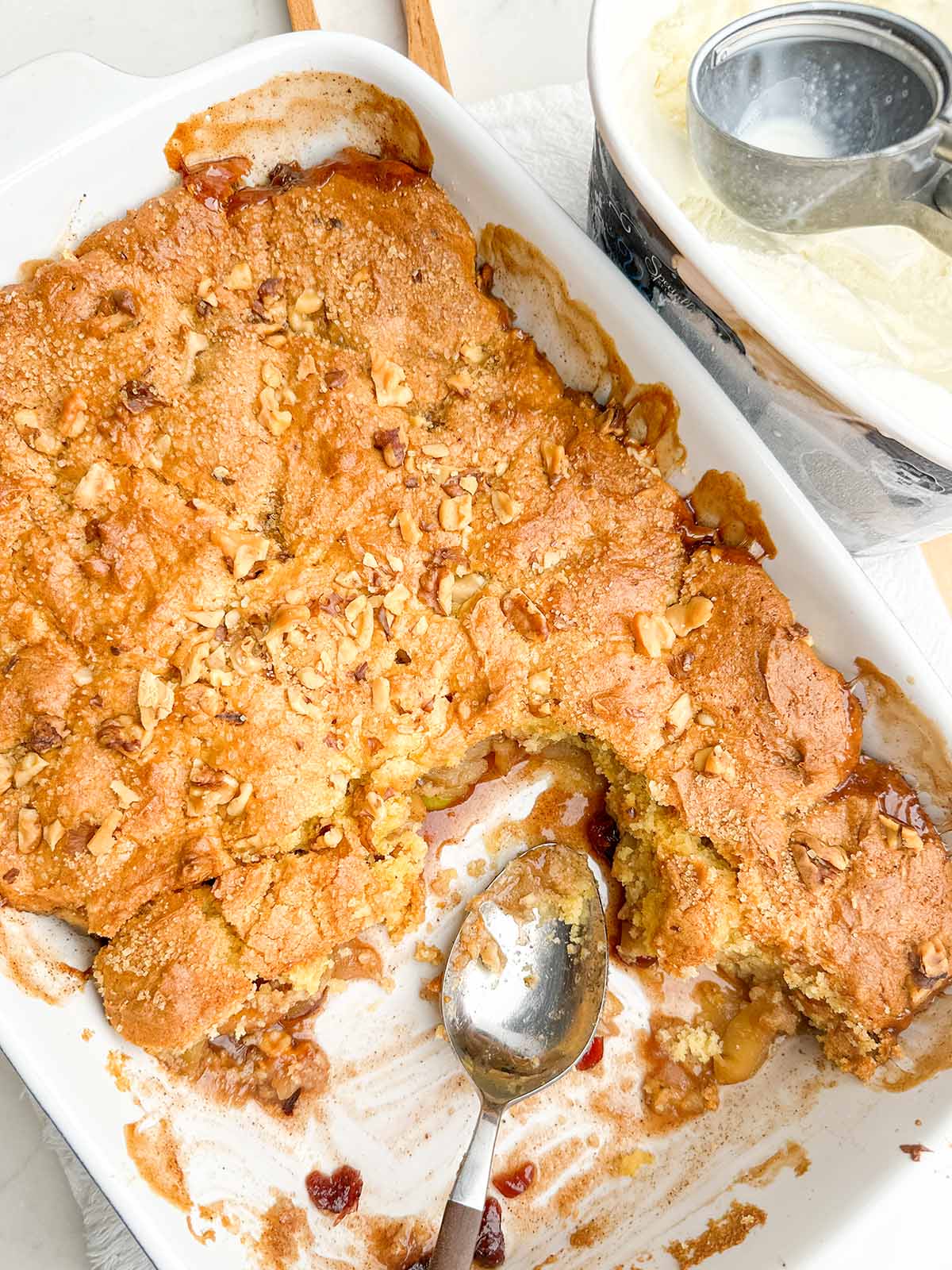 partial apple cobbler in a white baking dish with a spoon resting in it.