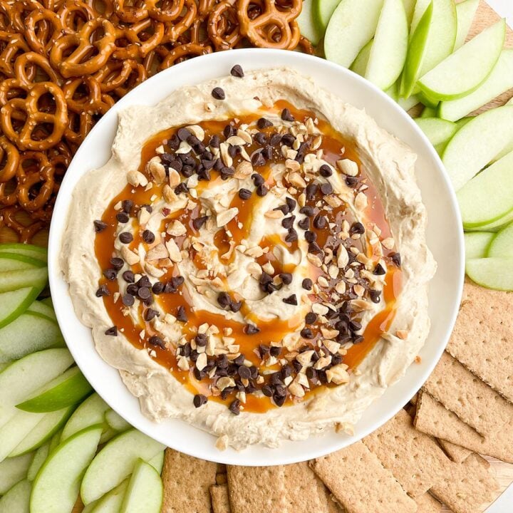 caramel apple dip in a white bowl on a round platter with pretzels, apples, and graham crackers
