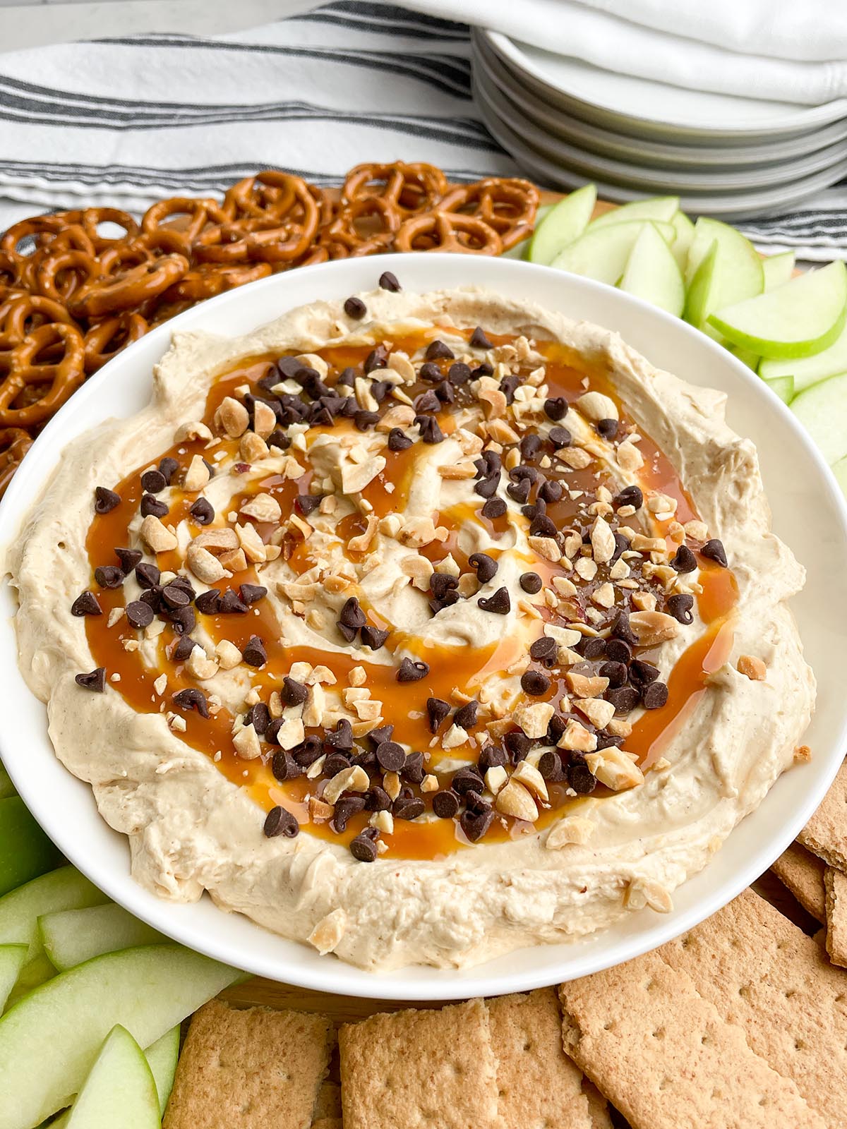 caramel apple dip in a white bowl on a round platter with pretzels, apples, and graham crackers