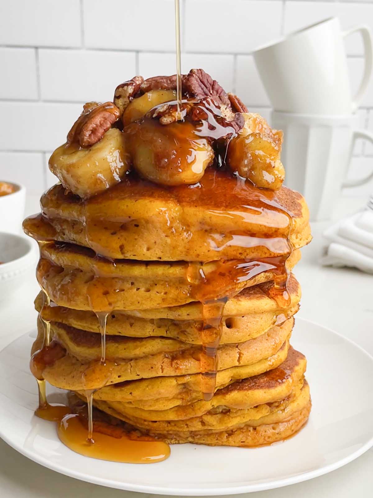 stack of pumpkin pancakes toped with caramelized bananas, toasted pecans, and syrup on a white plate.