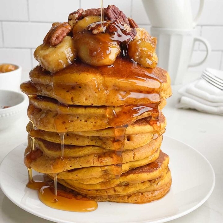 stack of pumpkin pancakes toped with caramlized bananas, toasted pecans, and syrup on a white plate