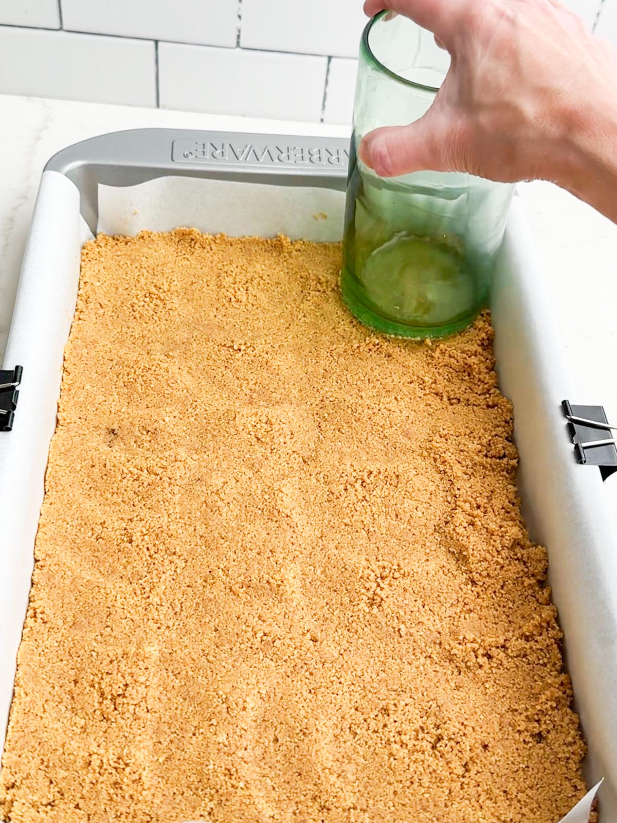 Hand holding a glass pressing Nutter Butter crumbs into a pan.