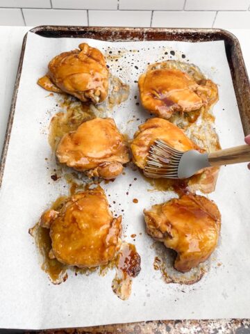 brush applying sauce to chicken thighs on parchment lined baking sheet.