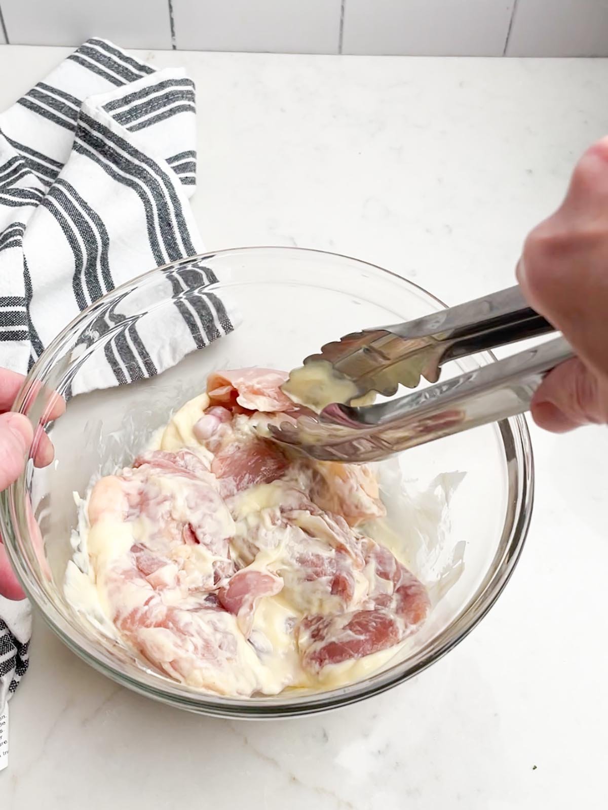 tongs turning chicken in mayo mixture in a clear bowl