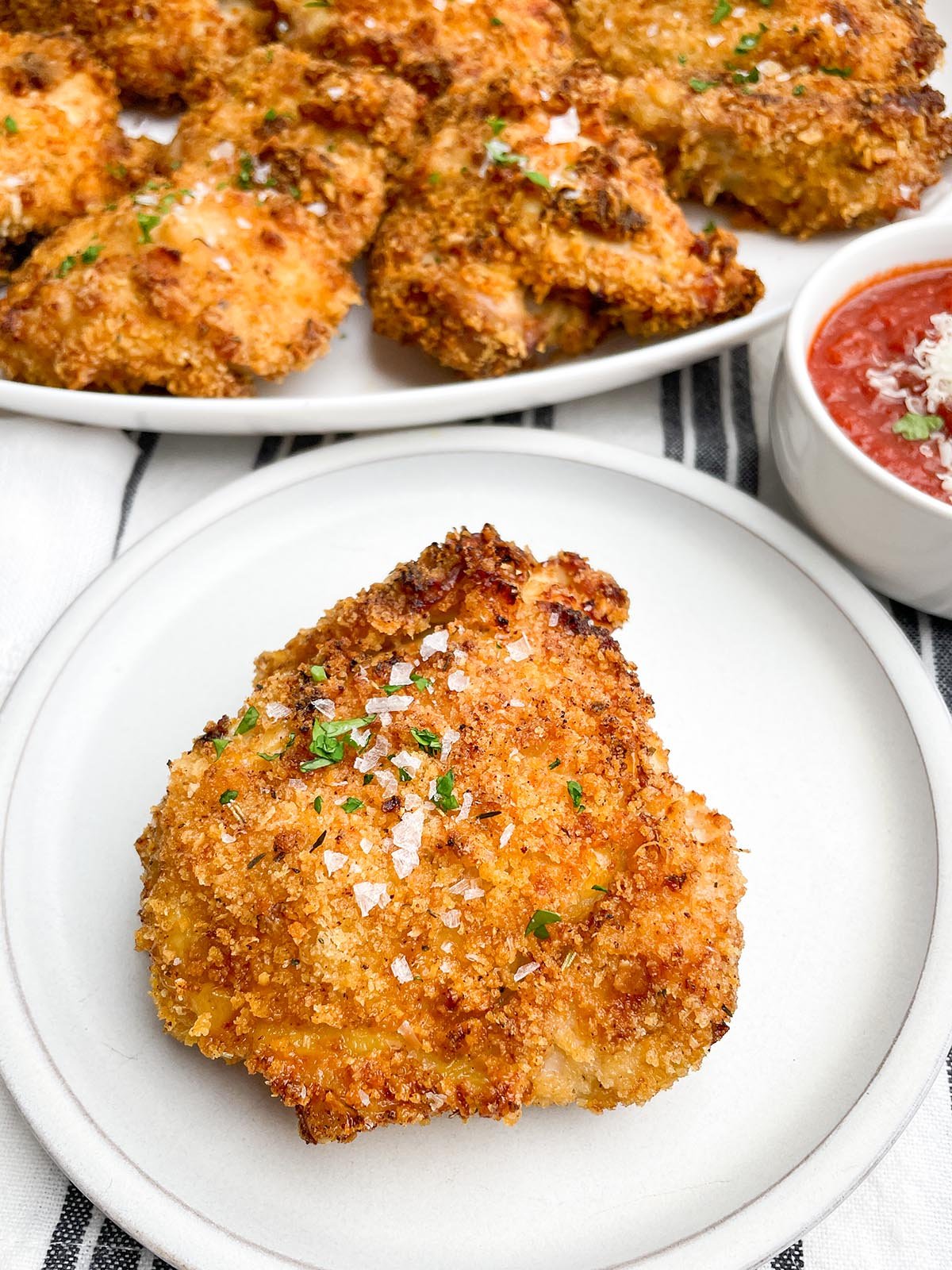 panko bone-in chicken thigh on a white plate with bowl of marinara and platter of chicken in background