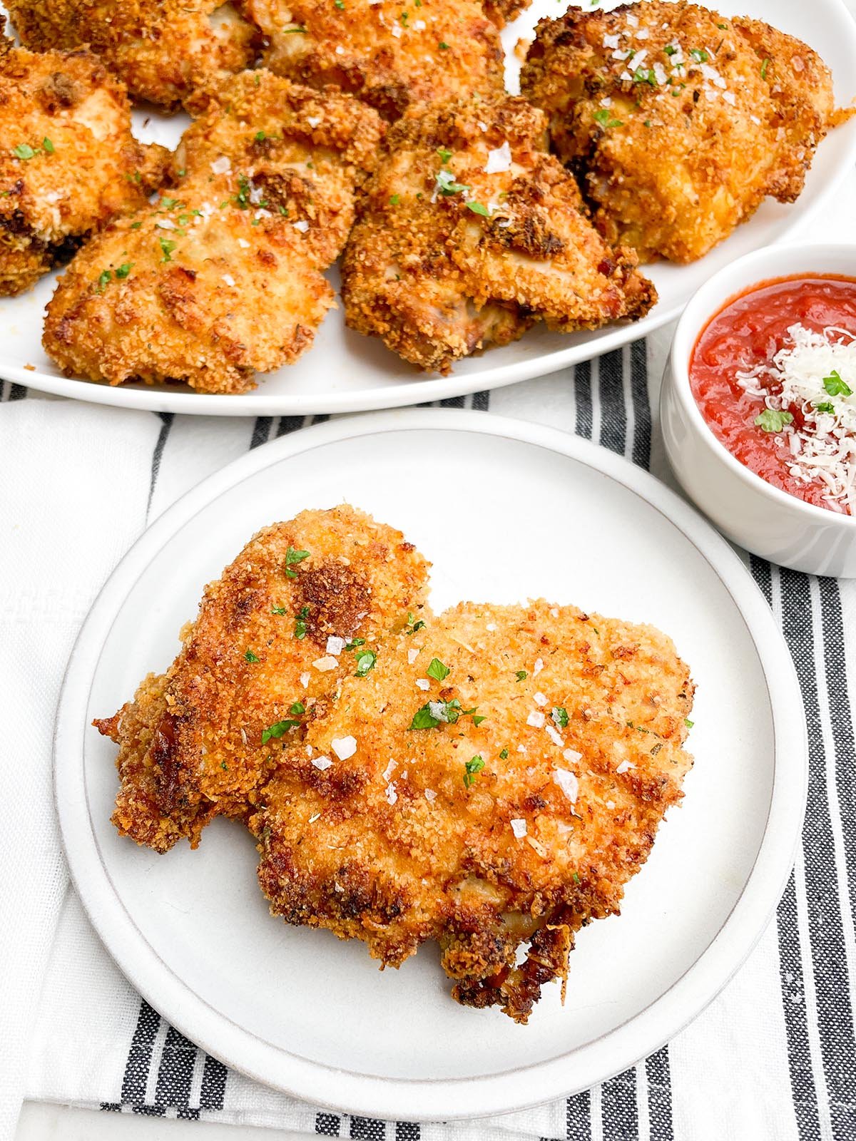 panko boneless chicken thigh on a white plate with bowl of marinara and platter of chicken in background