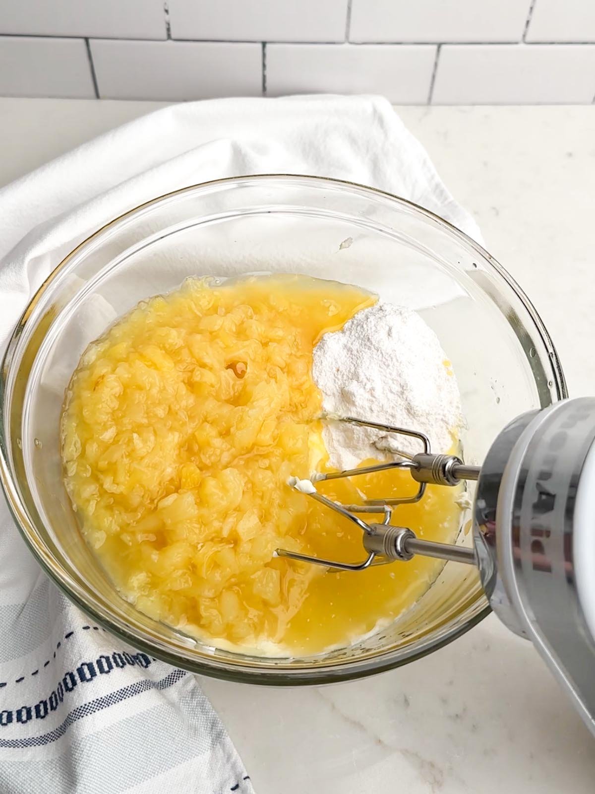 cream cheese, pudding mix, and crushed pineapple in a clear bowl with an electric mixer in it