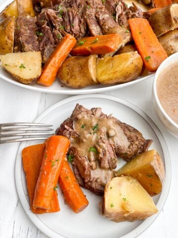 Pot roast with onion soup mix, potatoes, and carrots on a white plate with platter of pot roast in background.