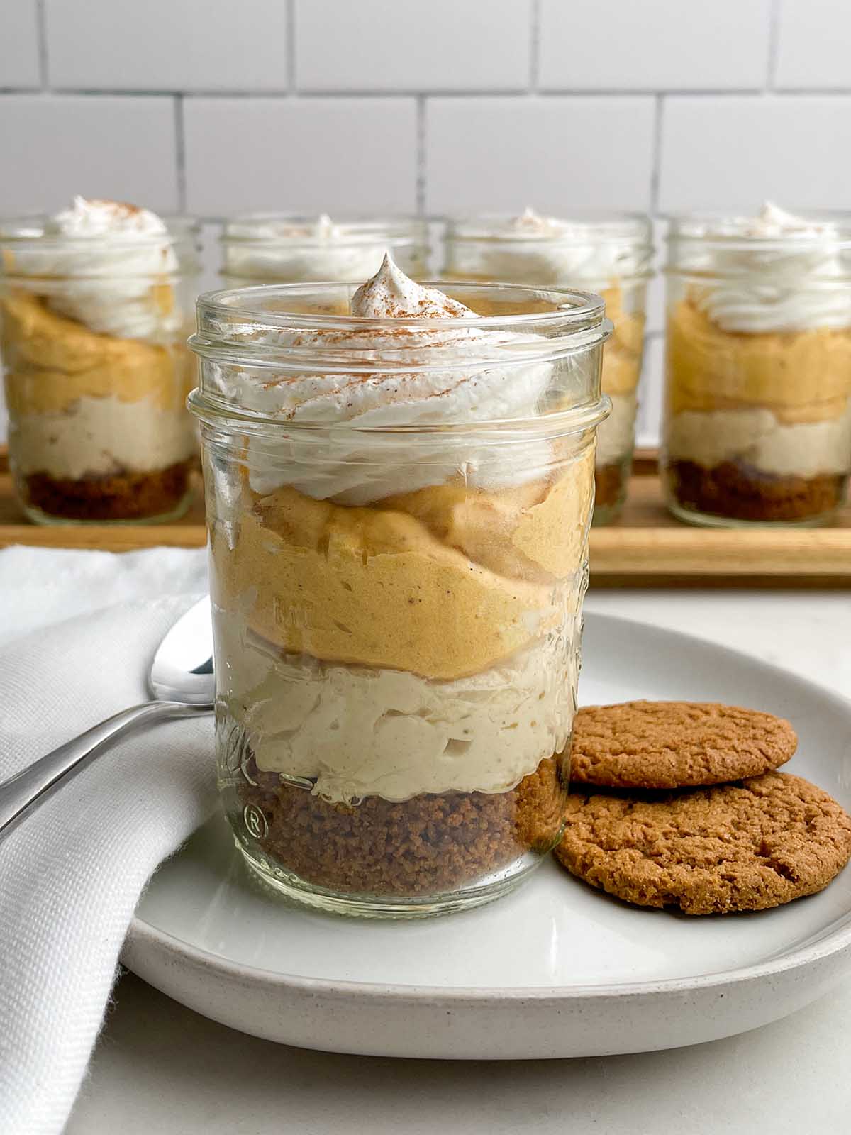 a pumpkin pie in a cup on a plate with gingersnap cookies, a napkin, and a spoon in front of jars of pumpkin pie in a cup