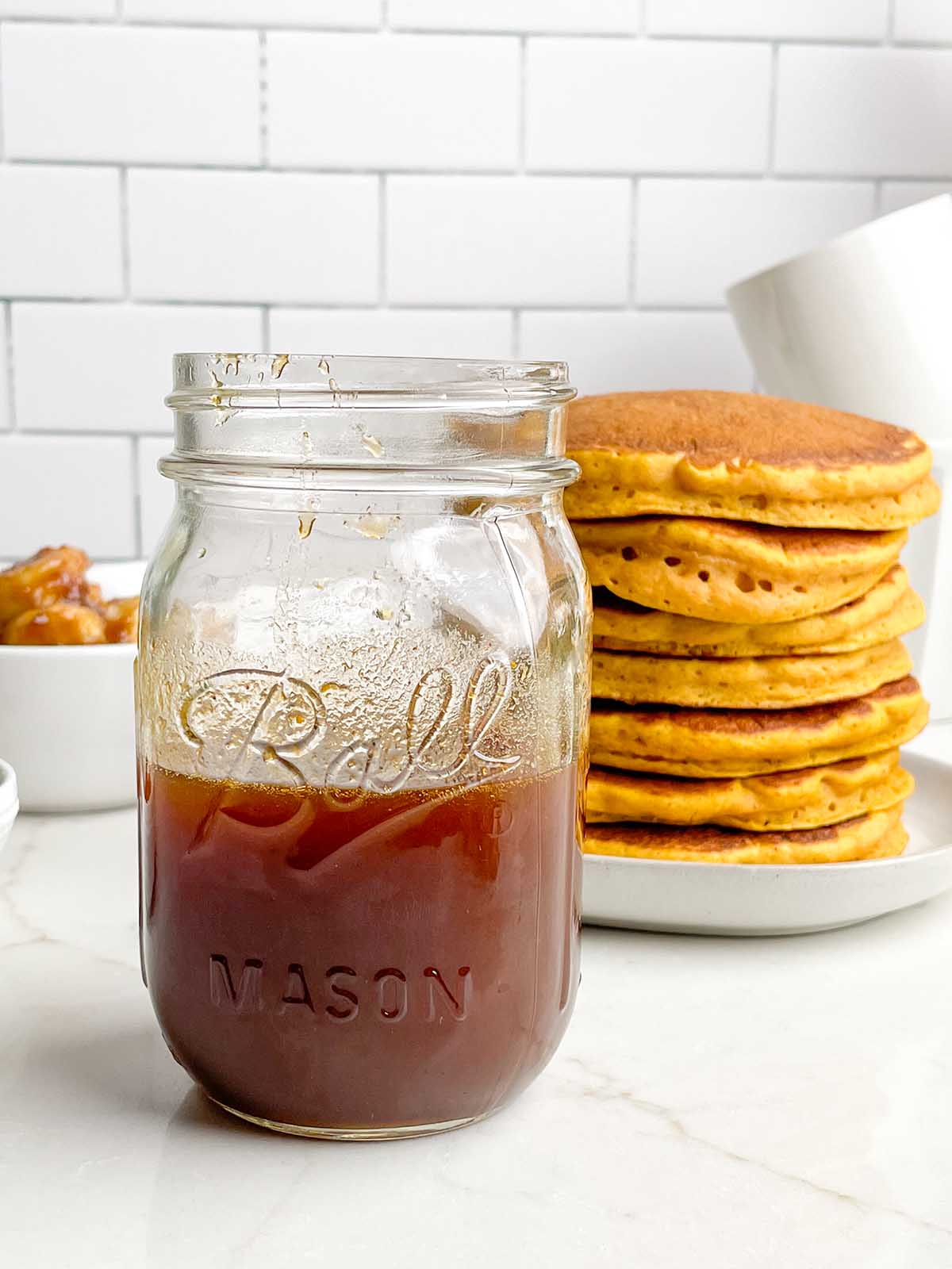 mason jar of pumpkin maple syrup in front of a stack of pancakes.