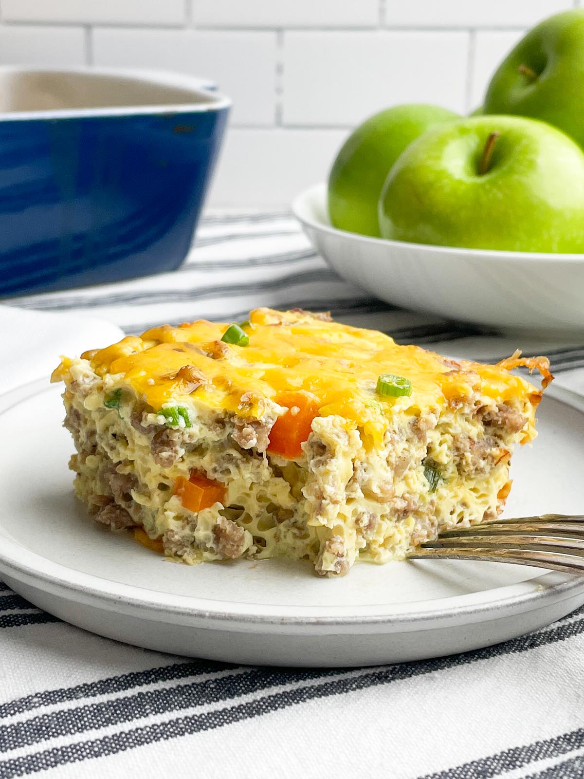 piece of sausage egg bake on a white plate with casserole dish and green apples in background.