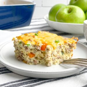 piece of sausage egg bake on a white plate with casserole dish and green apples in background