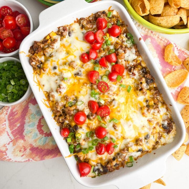 taco dip recipe with ground beef in white casserole dish with colorful napkin, plates, and scoops chips on counter