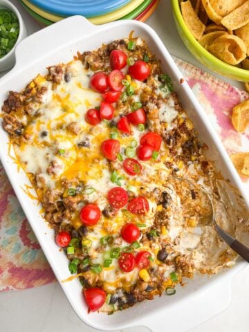 taco dip recipe with ground beef in white casserole dish with colorful napkin, plates, and scoops chips on counter.