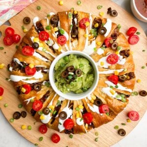 taco ring covered in toppings with guacamole in the center on a wooden cutting board