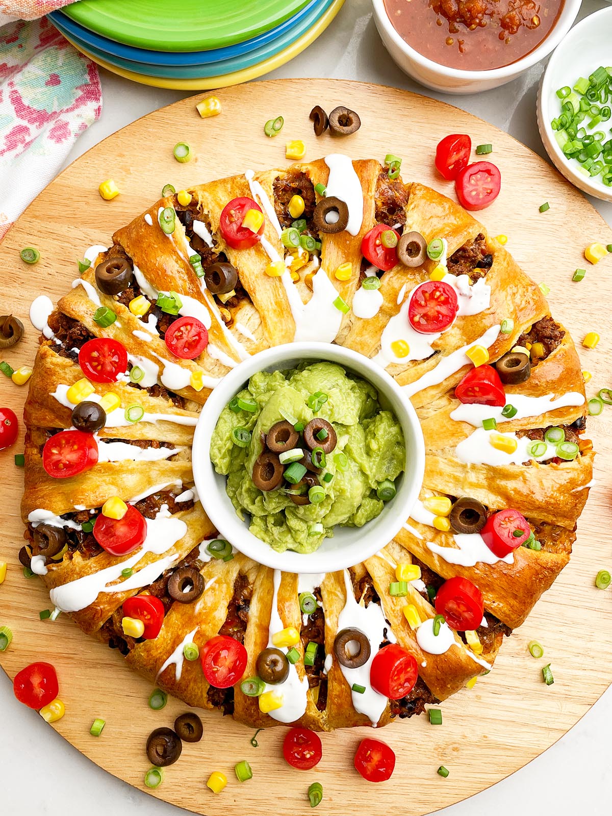 taco ring covered in toppings with guacamole in the center on a wooden cutting board