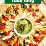 taco ring covered in toppings with guacamole in the center on a wooden cutting board